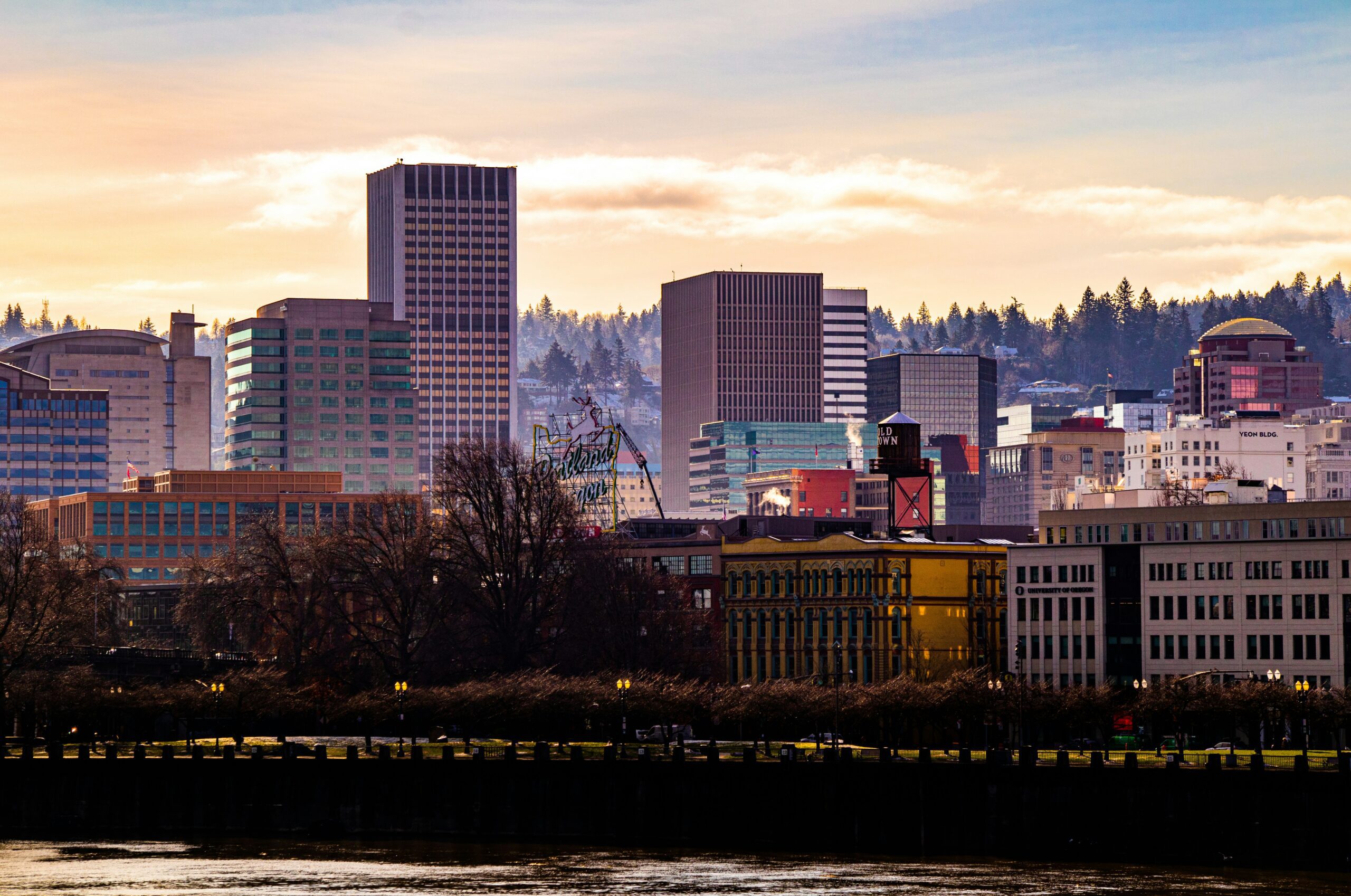 City skyline at sunset with buildings and trees along the waterfront.